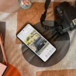 Điện thoại Android hiển thị ứng dụng Photoshop Beta trên giường, cạnh máy ảnh và sổ tay, minh họa khả năng chỉnh sửa ảnh di động.